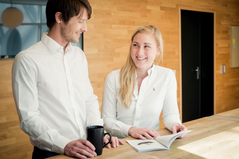 To kollegaer, en mann og en kvinne, i en vennlig samtale over en kaffekopp og en åpen bok på et lyst kontor med trepaneler i bakgrunnen